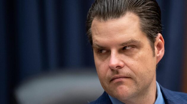 Matt Gaetz with combed-back hair and a suit gives a sideways glance, as if foreseeing the implosion of events backstage, all against a dark curtain background.