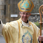 Amid the whispers of a scandal in the cathedral, the Archbishop of Canterbury, adorned in ceremonial attire and holding a staff, waves and smiles outdoors.